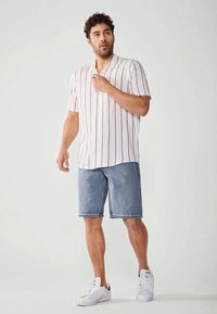 Camicetta bianca a maniche corte con righe verticali marroni abbinata a pantaloni shorts di denim, completata da sneakers bianche. Outfit estivo casual.