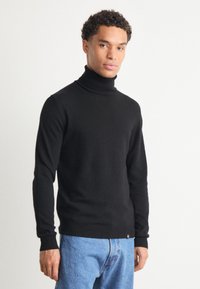 Μαύρο πουλόβερ φούτερ από λείο ύφασμα jersey; εφαρμοστό σχέδιο με μακριά μανίκια και ριμπ μανσέτες; φορεμένο με μπλε τζιν παντελόνι.