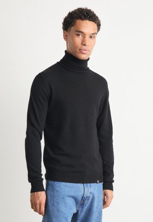 Maglione a collo alto nero in morbido tessuto a maglia; design aderente con maniche lunghe e polsini a coste; indossato con jeans in denim blu.