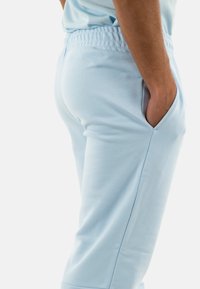 Pantaloni sportivi azzurri chiari con vita elastica, tasche laterali e una superficie liscia e morbida. Design aderente, mostrati di lato.
