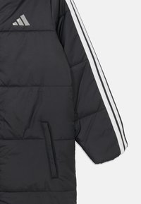 Mustan pehmustetun takin hiha, jossa on valkoiset kolmoisraidat ja pieni Adidas-logo olkapään lähellä, varustettuna vetoketjullisella sivutaskulla.