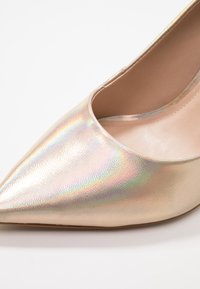 Metalické zlaté lodičky na vysokém podpatku s špičatou špičkou a hladkým povrchem, s iridescentním leskem a vnitřním polstrováním.