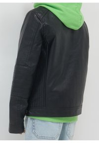 Czarna kurtka z sztucznej skóry z żebrowanym wykończeniem na rękawach, noszona na zielonym hoodie. Czyste linie, zorganizowany kształt i minimalistyczny design.