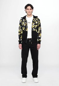 Μαύρη φούτερ ζακέτα με κίτρινο λουλούδι εμπριμέ πάνω από λευκό μπλουζάκι, συνδυασμένη με μαύρα τζιν και λευκά αθλητικά παπούτσια. Μοντέλο σε όρθια στάση.