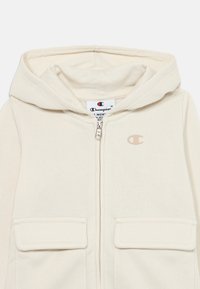 Krämfärgad hoodie med dragkedja fram, två framfickor och en liten logotyp på vänster bröst. Mjuk textur och avslappnad design.