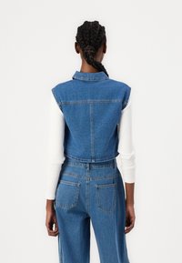 Denim brezrokavnik s ovratnikom, modri material, neobdelani robovi in stranskima žepoma, v kombinaciji z belim rebrastim dolgim rokavom.