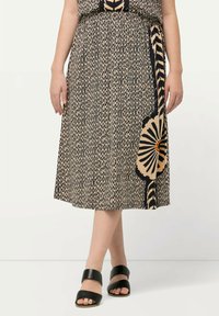 Jupe noire et beige à motifs avec une taille élastique, présentant un design floral et une fente sur le côté. Portée avec des sandales à talons noires.