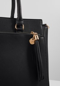 Borsa a tracolla in ecopelle nera con hardware dorato, caratterizzata da un accento con nappina e un'etichetta in ottone. Texture liscia e design strutturato.