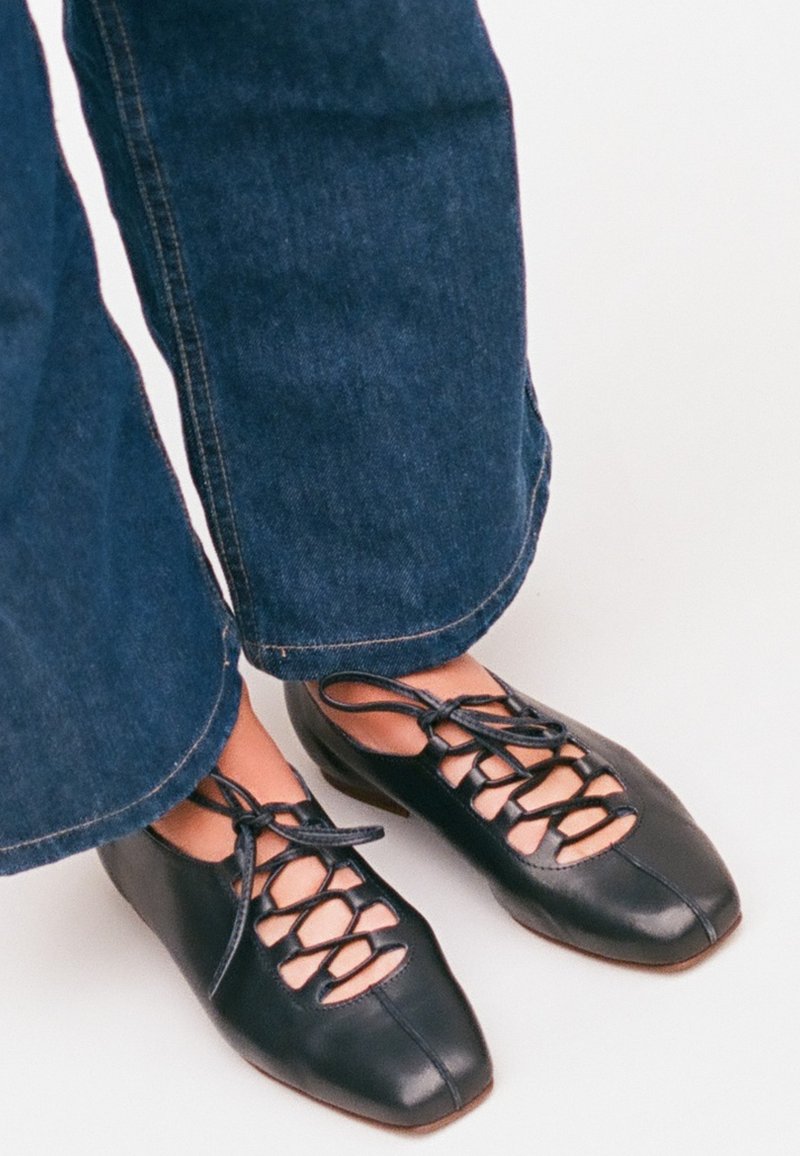 Nohy v tmavě modrých džínách obuté v černých kožených botách na šněrování s otevřenými výřezy na světlém pozadí.