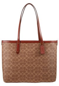 Sac fourre-tout marron avec monogramme, bord en cuir et doubles bretelles, présentant une étiquette en cuir attachée sur fond blanc.