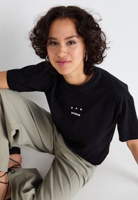 Zwarte katoenen t-shirt met "CPH STUDIOS" in het wit gedrukt, gecombineerd met lichtbeige wijde pantalons, getoond tegen een effen achtergrond.