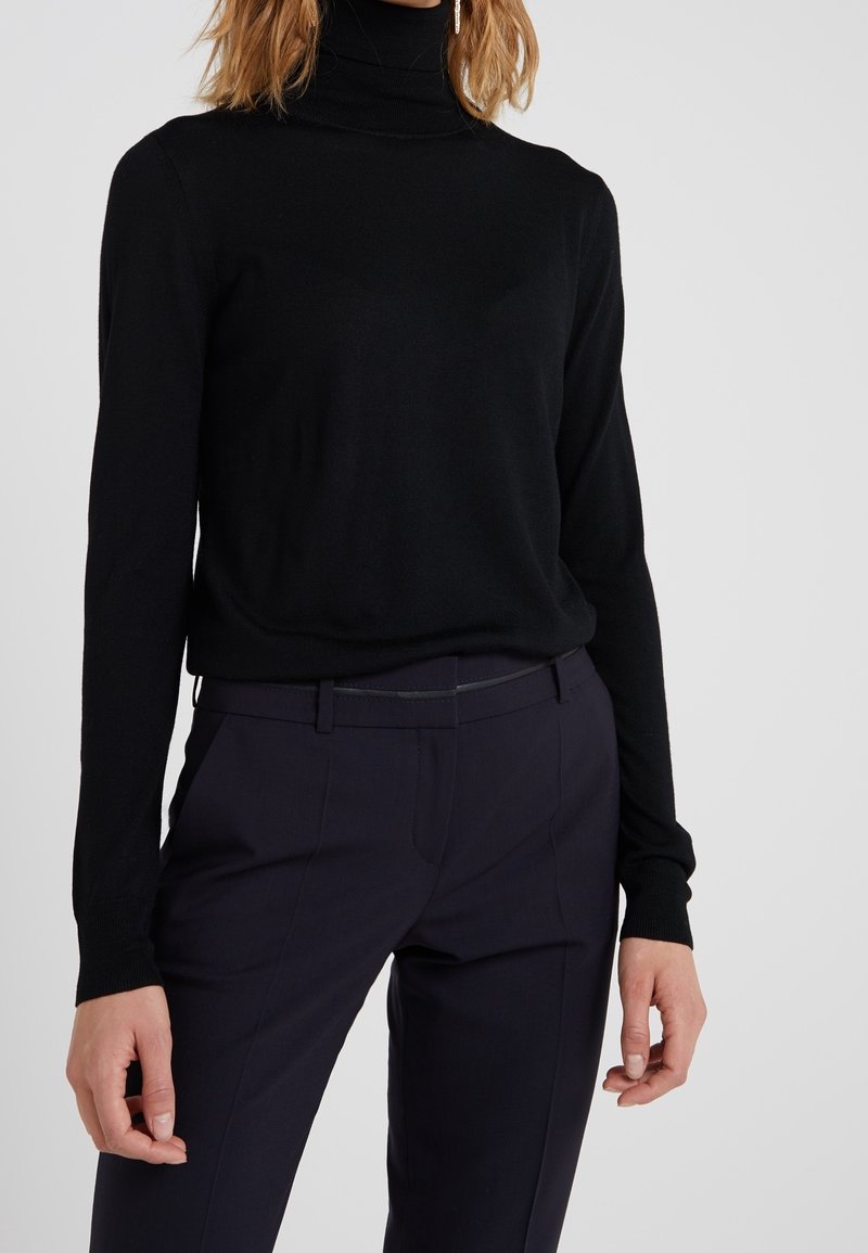 Pull à col roulé noir en tissu finement tricoté, avec manches longues et coupe ajustée, associé à un pantalon sombre et tailleur.