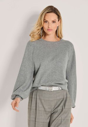 Vrouw met een lichtgrijze gebreide trui met ballonmouwen, geruite hoge taille broek, zilveren riem en lange hangende oorbellen.