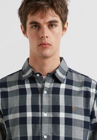 Marineblaue und graue karierte Hemd mit Spread-Kragen, ausgestattet mit weißen Knöpfen und einem kleinen orangefarbenen Logo auf der Brust.