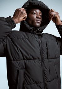 Jachetă puf din material negru, cu glugă, având închidere cu fermoar, manșete elastice și un finisaj texturat. Include buzunare laterale și un design ușor matlasat.