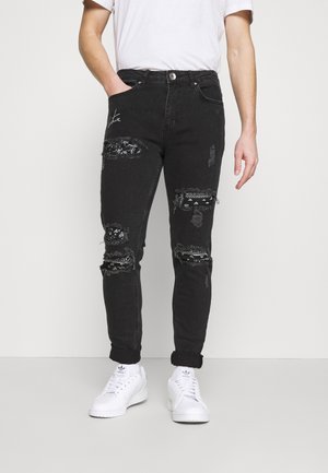 Mand iført sorte slidte jeans med mønstrede lapper og hvide sneakers, stående mod en ensfarvet lys baggrund.