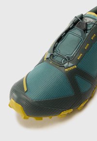 Teal and black trail running shoe with a textured mesh upper, yellow accents, a secure lacing system, and a rugged, grooved rubber sole.
