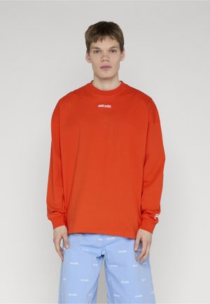 Joven con una camiseta de manga larga holgada de color naranja brillante con texto blanco pequeño y pantalones azul claro con patrones de texto blanco repetidos.