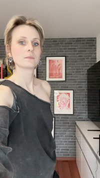 Crni top s otvorenim ramenima, s rubovima koji se trgnu, mekane teksture. Pozadina sa sivim zidom od opeke i uokvirenom roza umjetničkom djelu. Minimalistički dekor.
