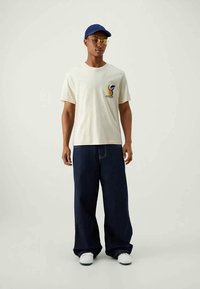 Homem a usar um boné azul, óculos com lente amarela, t-shirt creme com gráfico, calças largas azul-escuro e ténis brancos a pousar contra um fundo simples.