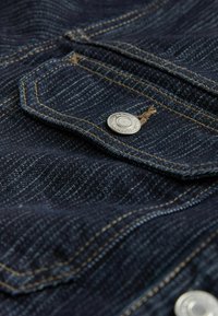 Mørkeblå denim stof med en sølvfarvet metaltrykknap, synlige stikninger i kontrastfarvet guld og en tekstureret overflade med subtile linjer.