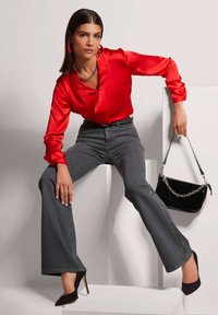 Rode satijnen blouse met lange mouwen, grijze hooggotige flared jeans, zwarte stilettos en een zwarte handtas met zilveren kettingaccenten.