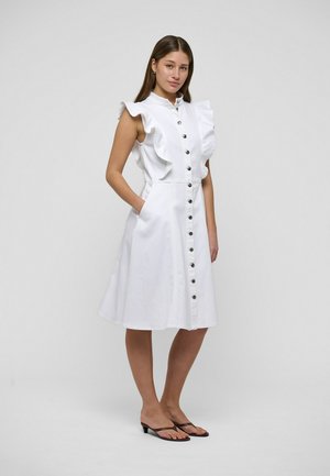 Jeune femme portant une robe midi blanche sans manches avec des épaules à volants et des boutons sur le devant, debout les mains dans les poches, sur un fond uni.