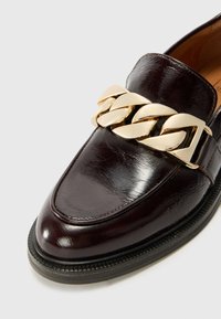 Dark brown leather loafer featuring a prominent gold chain accent across the vamp, smooth texture, and a structured shape with a raised heel.