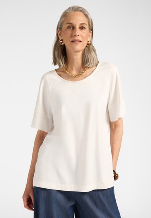 Frau mit grauen Haaren, die ein weißes kurzärmeliges Oberteil, blaue Jeans, eine goldene Halskette, goldene Ohrringe und ein braunes Armband trägt und vor einem einfarbigen Hintergrund posiert.