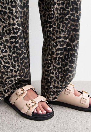 Sandales beiges avec des boucles dorées, semelles noires et design ouvert, associées à un pantalon à imprimé léopard noir et beige.