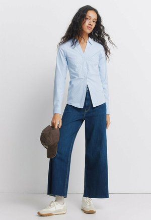 Nainen, joka pukeutunut vaaleansiniseen, raidalliseen nappipaitaan, leveälahkeisiin tummansinisin farkkuihin ja valkoisiin lenkkareihin, pitää toisessa kädessään ruskeaa baseball-lippistä.