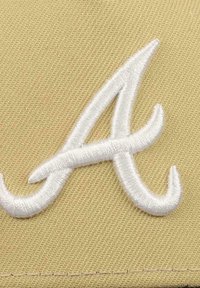 Tan baseball cap featuring a prominent white embroidered "A" logo. The fabric has a textured finish with visible stitching.