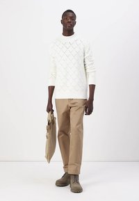 Maglione bianco a maglia con motivi a rombi, pantaloni beige e stivaletti grigi alla caviglia. Tiene una borsa a tracolla color cammello. Sfondo semplice.