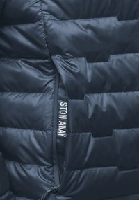 Marineblaue Steppjacke mit einem gesteppte Design, einer Reißverschlusstasche und "STOW AWAY" auf dem Reißverschluss bedruckt. Glatte, glänzende Textur.