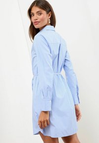 Abito camicia a righe blu e bianche con maniche lunghe, polsini con bottoni, colletto e vita aderente, dotato di uno spacco posteriore per facilitare i movimenti.