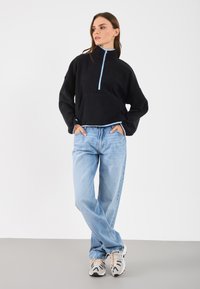 Pulover scurt din fâscă neagră, cu guler înalt, fermoar albastru și buzunar frontal; asortat cu blugi albaștri deschiși și adidași cu model.