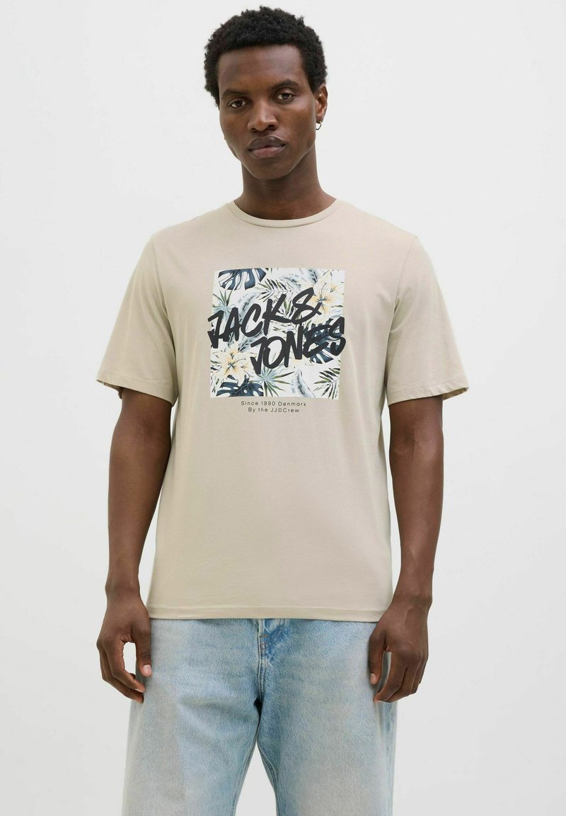 Camiseta de color beige claro con cuello redondo, que presenta un estampado gráfico de hojas tropicales y el texto "JACK & JONES" en letras negritas negras.
