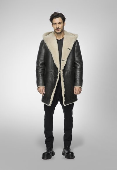 Cappotto di pelle nera con colletto e fodera in shearling beige, caratterizzato da un design doppiopetto, indossato sopra pantaloni e scarpe nere.