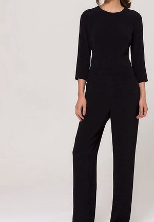 Woman wearing a fitted black jumpsuit with three-quarter sleeves and wide-leg pants, standing against a plain light background.