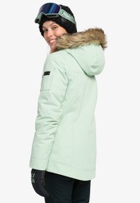 Mint green ski jacket with a fur-lined hood, zippered side pocket, and a sleek design. Paired with black gloves for functionality.