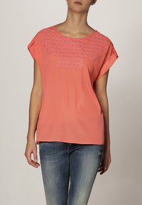 Koral kortærmet bluse med et tekstureret broderet mønster på brystet, lavet af letvægtsstof, parret med blå jeans.