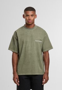 Mladý muž s krátkými vlasy, oblečený v nadměrném olivově zeleném tričku s nápisem "Sunday Running Club" a černých šortkách na jednobarevném pozadí.