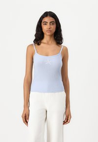 Light blue ribbed camisole with adjustable spaghetti straps, featuring a central star-shaped embroidery. Worn with white wide-leg pants.