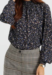 Blommig blus i mörkblått, krämfärgat och brunt, med rynkad halsringning och puffärmar, ihopparad med rutiga högmidjade byxor.