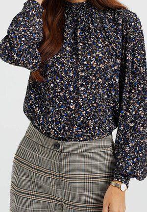 Blusa floreale in blu scuro, crema e marrone, con scollatura arricciata e maniche a palloncino, abbinata a pantaloni a quadri a vita alta.
