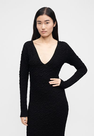 Frau mit langem schwarzen Haar, die ein strukturiertes schwarzes langärmliges Kleid mit tiefem V-Ausschnitt trägt und mit einer Hand in der Hüfte vor einem weißen Hintergrund posiert.