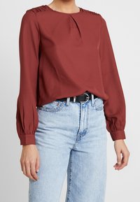 Rustinvärinen paita, jossa on laskokset hartioilla, pitkät hihat ja cuffit, yhdistettynä korkeavyötäröisiin vaaleansinihin farkkuihin.