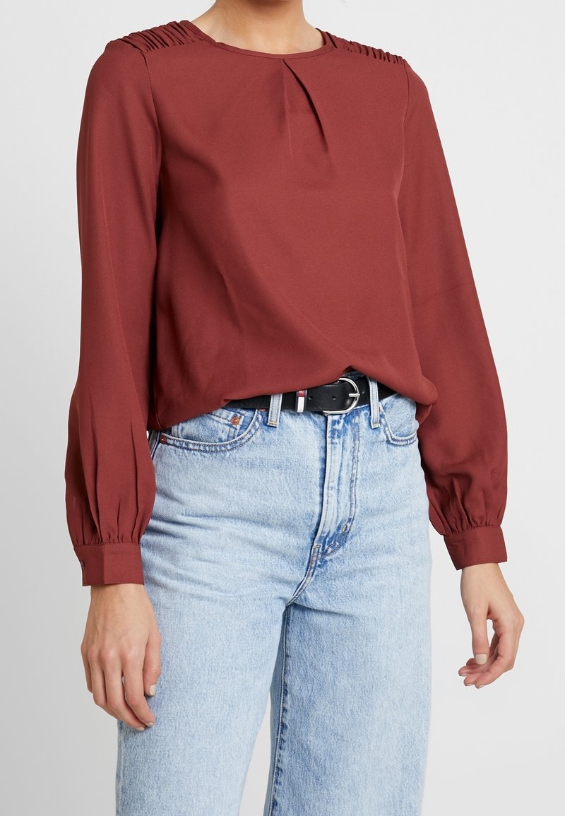 Rustinvärinen paita, jossa on laskokset hartioilla, pitkät hihat ja cuffit, yhdistettynä korkeavyötäröisiin vaaleansinihin farkkuihin.