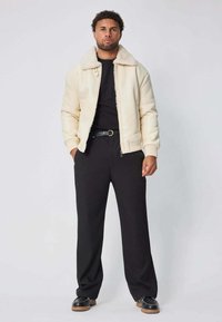 Blouson créme avec un col en fausse fourrure, t-shirt noir en dessous, assorti à un pantalon noir ample et des chaussures noires avec une semelle en caoutchouc.