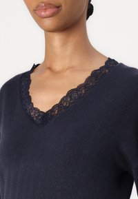 Kvinna som bär en marinblå V-ringad tröja med spetsdetaljer längs halsringningen, i stil med att visa halsen och övre bröstområdet.